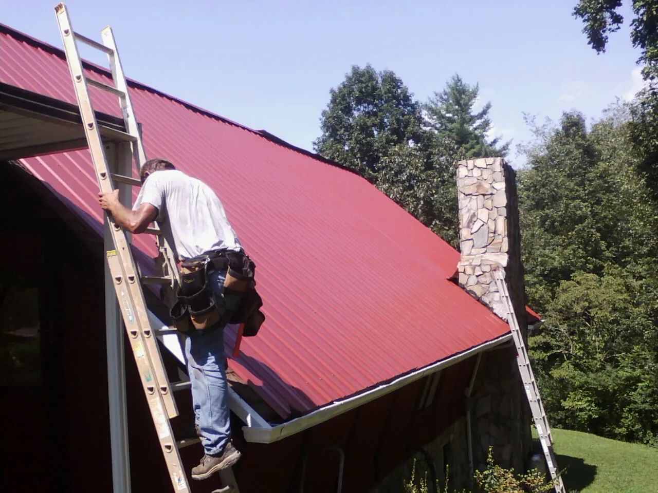 Roofer on a ladder beside a red metal roof and stone chimney — Soffit & Fascia Repair by Roofing Brothers in Allenport